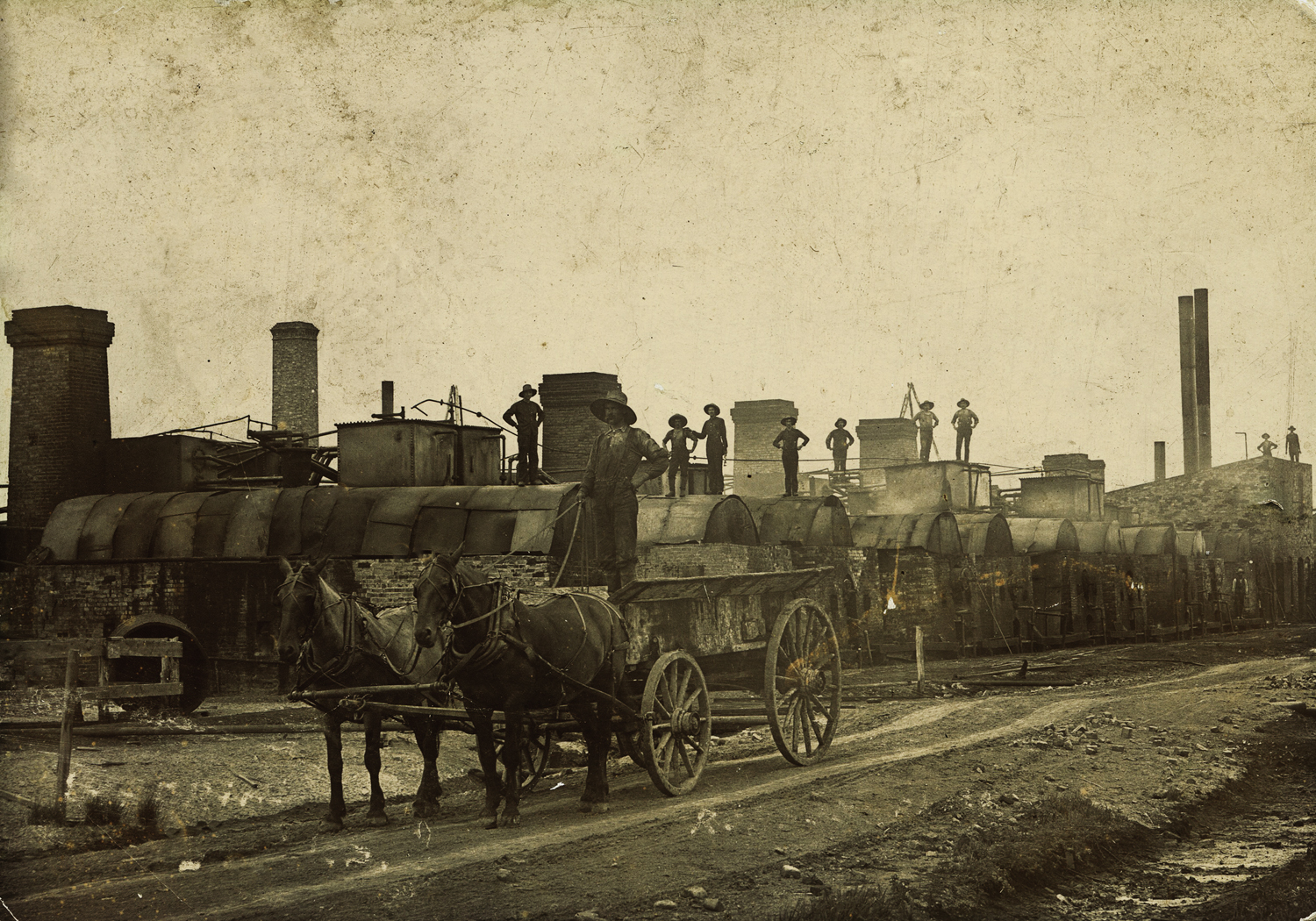An image showing an early oil refinery, with muddy roads, a man standing on a wagon and several oil workers posing on the tops of refining equipment. 