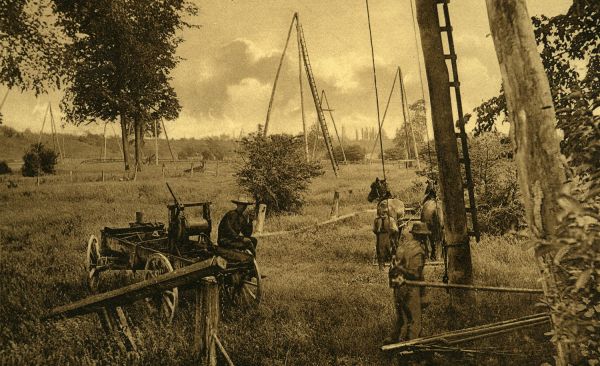 A photo of a field showing oil equipment including three pole derricks and jerker lines. There is a man sitting on a wagon. 