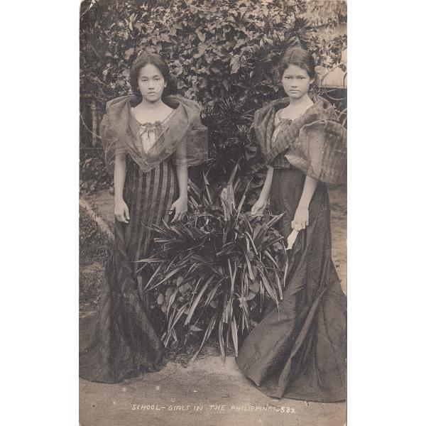 Devant d'une carte postale illustrant deux écolières vêtues de jolies robes et se tenant de chaque côté d'un arbuste hérissé avec des arbres en arrière-plan. Les ourlets des robes touchent le sol et les jeunes filles portent des vêtements transparents sur leurs épaules. Leurs cheveux sont séparés et attachés. 