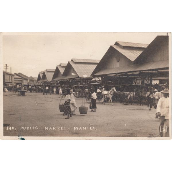 Front of a postcard showing three identical wooden buildings with two peaks in the roof. There is a line of carts in front of them. There are people walking around in an open space and a man is carrying two baskets connected by a pole on his shoulder. 