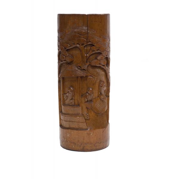 The front of a brown carved wooden vase depicting three men near a structure with a roof and a tree in behind. 