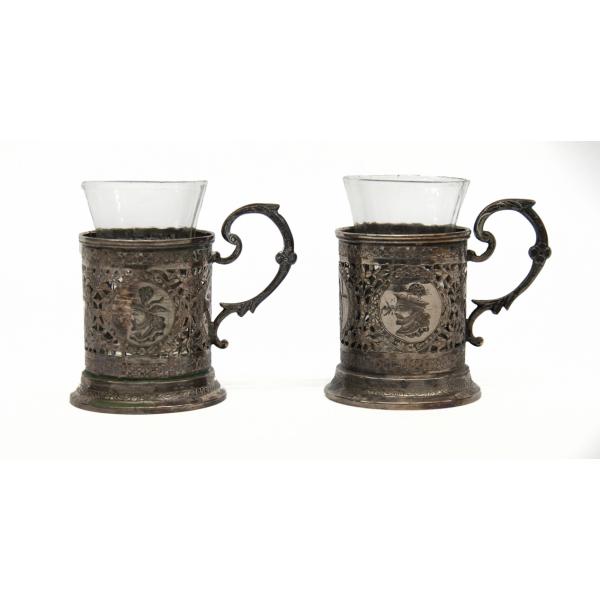 Two tea cups side by side with their glass inserts. Each has the profile of a king's head and a floral design. The handles on the cups are curved. 