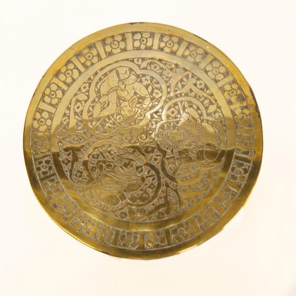 The top of a brass pyxis with three kneeling figures surrounded by a floral design. 