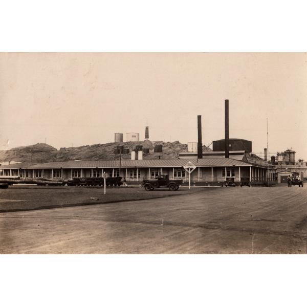 A long one-story building with white pillars on the porch. There is a wide road in front and a railway crossing. Carts are in front of the building and there are four cars in the photo. There are storage tanks on the hill in the background. 