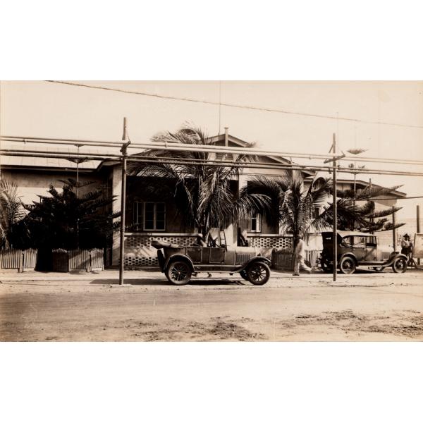 Photo d'un bâtiment d'un étage avec des treillis sur le porche et des palmiers à l'avant. Deux voitures sont stationnées à l'extérieur, le long d'un chemin de terre. Des fils électriques passent au-dessus.