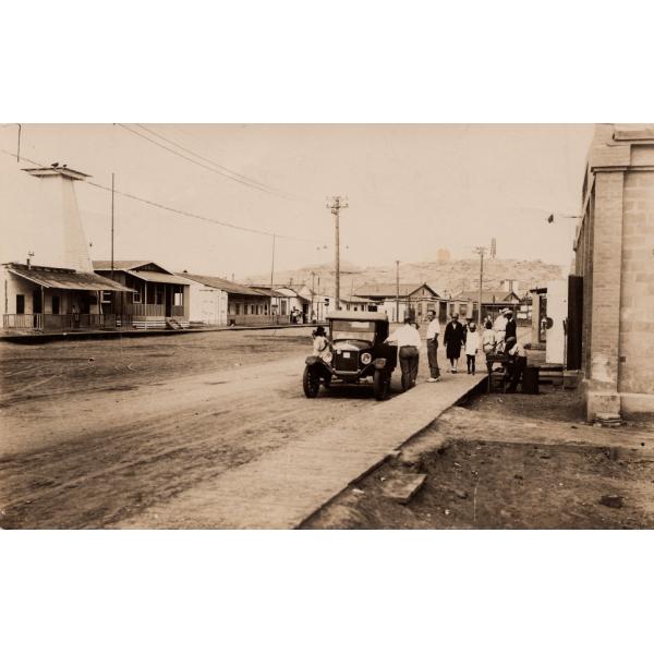 A photo of a dirt road with a wooden sidewalk running beside it. There is a group of people near a car parked on the side. There are single-story buildings on either side and hills in the background. 