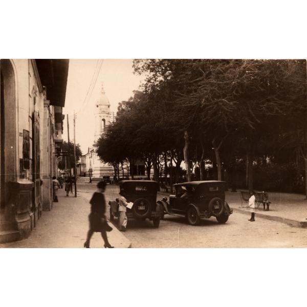 Une photo de deux voitures stationnées côte à côte sur une rue pavée. Il y a un garçon de chaque côté des deux voitures et un bâtiment à gauche avec une femme qui passe le coin en marchant. Un groupe d'arbres se trouve à droite et on aperçoit une tour blanche en bas de la route.