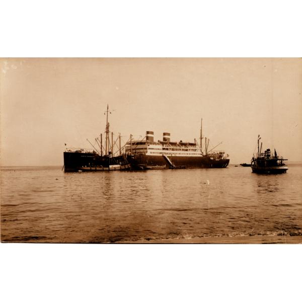 A photo of a ship with two masts and two smoke stacks, anchored in the water. There is a smaller boat just off to the left. There are people getting into small boats at the base of the large boat. 