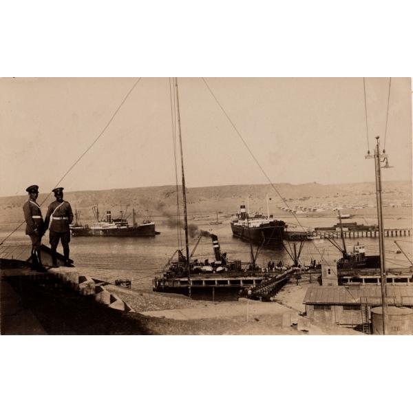 A photo of two men in uniform standing on a hill that leads down to a harbour where three large boats are docked. There are hills in the background. 