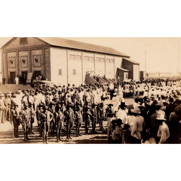 A photo of a crowd of people watching a group of men in uniform. There is a large building with three main doors in behind, as well as a car at the base of a set of steps. 