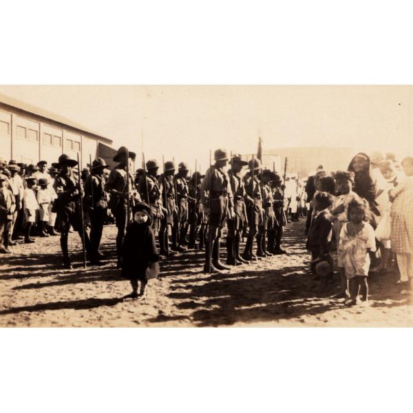 Une photo d'une foule regardant trois rangées d'hommes en uniforme, devant un grand bâtiment. Un jeune enfant regarde l'appareil photo et se tient devant la deuxième rangée d'hommes en uniforme. 