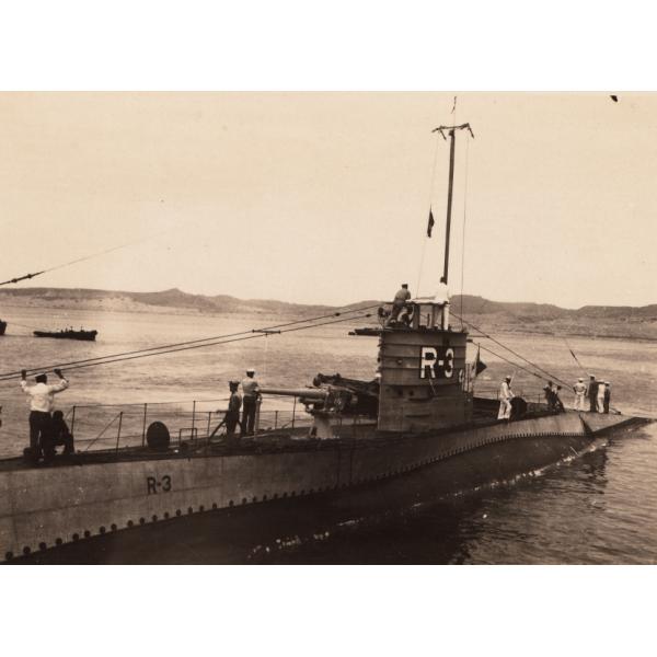 A photo of twelve men standing on the top of a submarine that is above water. It has "R-3" written on it. There is a small boat in behind it and hills in the background.  