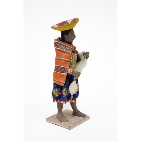 Side view of a figurine of a woman wearing sandals and who is dressed in a blue skirt with an embroidered design along the edge, as well as an orange rectangular shoulder cloth fastened at the front. She has an inverted yellow hat and is carrying a spool and wool. 