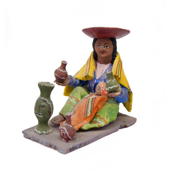 A figurine depicting a woman wearing a green skirt, a purple blouse, a blue vest with a design on it, and a yellow rectangular shoulder cloth. She is wearing an inverted red hat. There is a baby in her lap and she is holding pieces of pottery. 