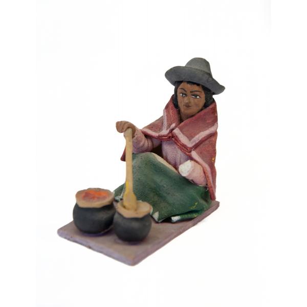 Figurine d’une femme remuant le contenu de deux marmites noires avec une cuillère brune. Elle porte des sandales, une jupe verte et un châle rectangulaire rouge. Un tablier blanc est enroulé autour de son poignet et elle porte un chapeau gris, avec le bord recourbé vers le haut. 