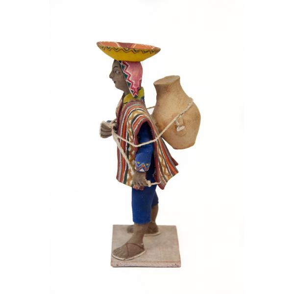 Side view of a figurine of a woman carrying a water jug on her back. She is wearing sandals, a blue suit, and a poncho of different colours, as well as a pink head covering and a yellow and orange upturned hat.