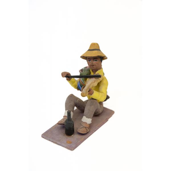 Figurine d’un homme jouant du violon et portant des sandales, des pantalons bruns à revers, une chemise jaune, ainsi qu’un sac vert noué sur une de ses épaules. Il porte un chapeau jaune et une bouteille noire est à ses pieds. 