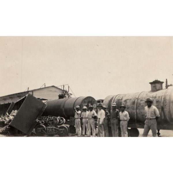 A photo of a group of men looking at a railway carriage that was derailed. There are two cylindrical carts still on the tracks and a building in behind. 