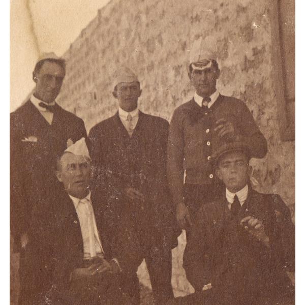 Cinq foreurs internationaux, trois debout et deux assis. Ils portent des costumes foncés. Quatre des hommes portent des chapeaux en papier et un (en bas à droite) porte un béret Ascot.  