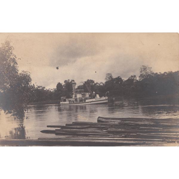 Front of postcard showing a white boat on a river. There is a pile of logs in the foreground and trees in the background.  