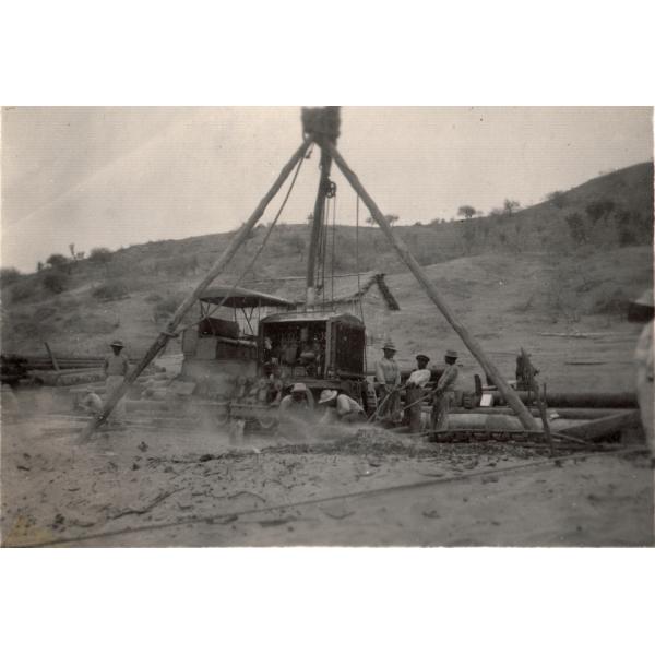 A group of men are putting together a bulldozer using three poles tied together at the top with a chain coming down from the middle to help lift the heavy pieces. There is a hill dotted with trees in the background. 