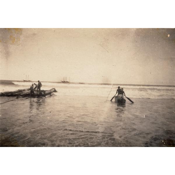 Three men standing on a raft made from logs and three men in a canoe. They are in shallow water and there are small waves moving towards them. There are two ships out on the water in the background.