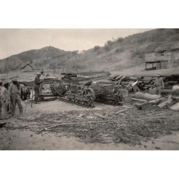 A photo of a half-assembled bulldozer. George Rawlings stands to the left. There are stacks of pipes in behind the bulldozer and two buildings on the hill at the back of the scene.