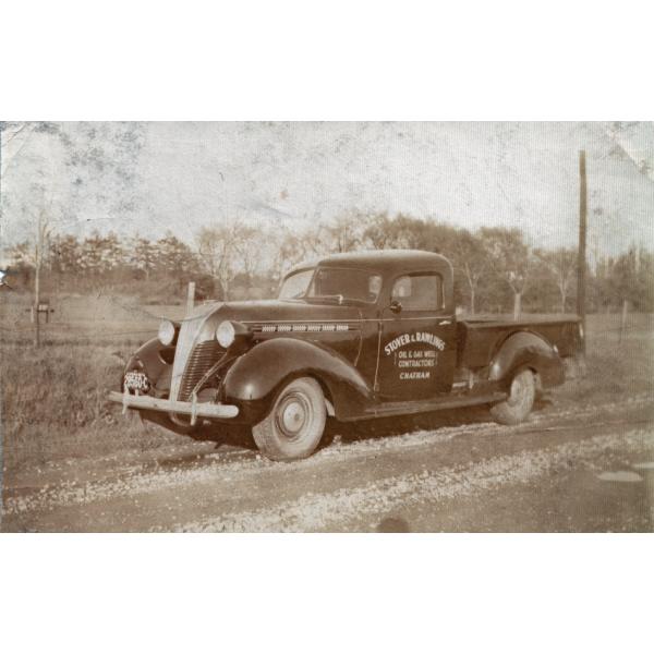 Un camion à deux portes est stationné sur le côté d’une route de terre, avec un champ et des arbres à côté de celui-ci. Le camion est foncé et « Stover and Rawlings. Oil & Gas Well Contractors. Chatham » est inscrit sur la porte. 