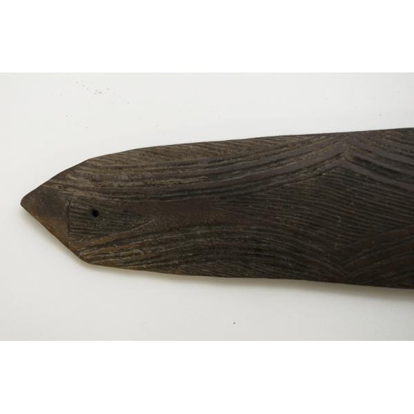 The tip of a dark wooden boomerang with a small hole in it. There is a line pattern carved into the wood.