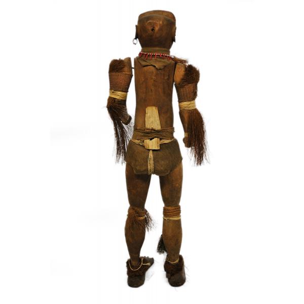 The back of a dark brown wooden doll with articulated arms and legs. It is decorated with tufts of grass around its neck, arms, knees, and ankles. There is a piece of cloth around its groin. It has a necklace and earring made from white and red beads