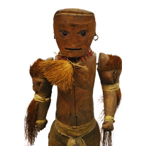 The upper half of a dark brown wooden doll. It is decorated with tufts of grass around its neck and arms. There is a piece of cloth around its groin. It has a necklace and earring made from white and red beads. Dark eyes, eyebrows, and a mouth have been carved into the face.