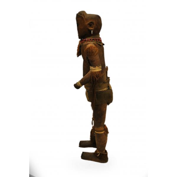 A dark brown wooden doll from the side. It is decorated with tufts of grass around its neck, arms, knees, and ankles. There is a piece of cloth around its groin. It has a necklace and earring made from white and red beads