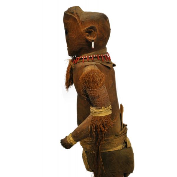 The side of the upper half of a dark brown wooden doll. It is decorated with tufts of grass around its neck and arms. There is a piece of cloth around its groin. It has a necklace and earring made from white and red beads. Dark eyes, eyebrows, and a mouth have been carved into the face.  