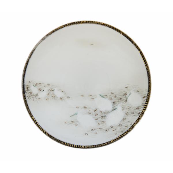 The top of a saucer decorated with a gold rim and seven white ibis birds nesting in a field with grass and gold dots.