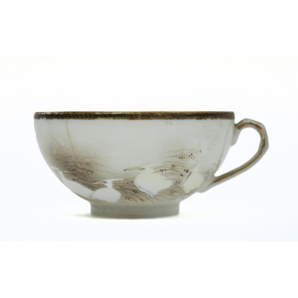 A tea cup with a gold rim, gold lines on the handle, and decorated with four white ibis birds nesting in a field of grass with gold dots.