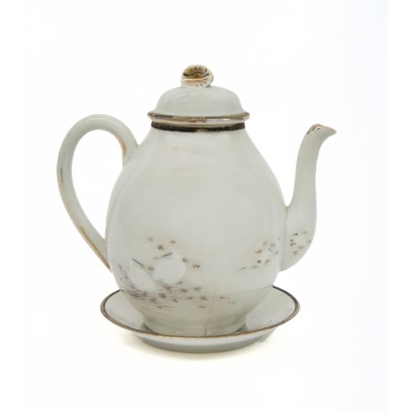 A white china teapot facing to the right, resting in a saucer, decorated with gold along the edges and with white ibis birds. The handle of the lid has a gold starburst.  