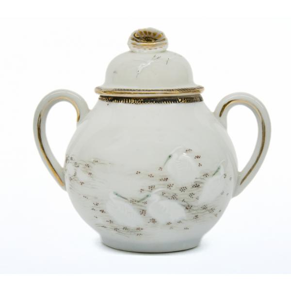 A white china sugar bowl decorated with gold rim and white ibis birds in a field with grass and gold dots. The lid has a gold starburst at the top and a gold rim at the bottom. 