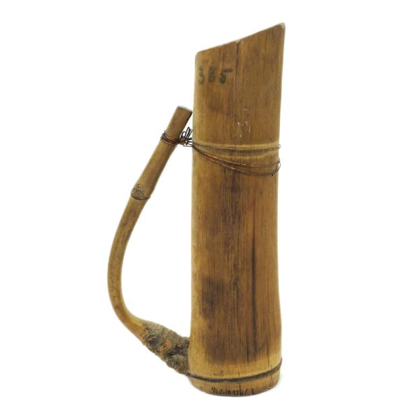 A light brown pitcher made from bamboo, with a handle facing left. The handle is made from a branch growing off of the trunk that was bent back and secured with copper wire.