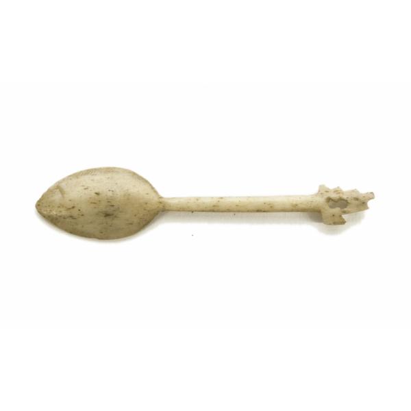 The back of an ivory-carved spoon with a point on the end. It is speckled with brown dots and the top of the handle is shaped like a bud. 