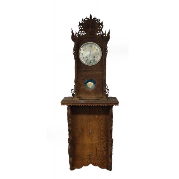 A brown wooden grandfather clock with a peacock carved into the top and carved dragons on the supports. There is a floral pattern around the clock face and pendulum case. The clock is off-white with gold numbers and hands. The pendulum is also off-white.