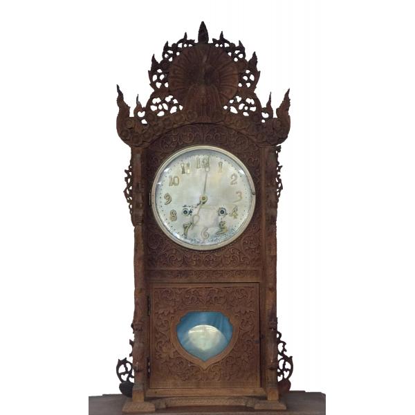The top section of a brown wooden grandfather clock with a peacock carved into the top. There is a floral pattern around the clock face and pendulum case. The clock is off-white with gold numbers and hands. The pendulum is also off-white.