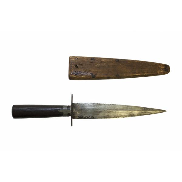 A metal dagger with a symmetrical blade and a wooden sheath, pointing to the right. The dagger has a dark wooden handle and two rows of dots along the blade. The brown wooden sheath has four nails on either side along the edge. 