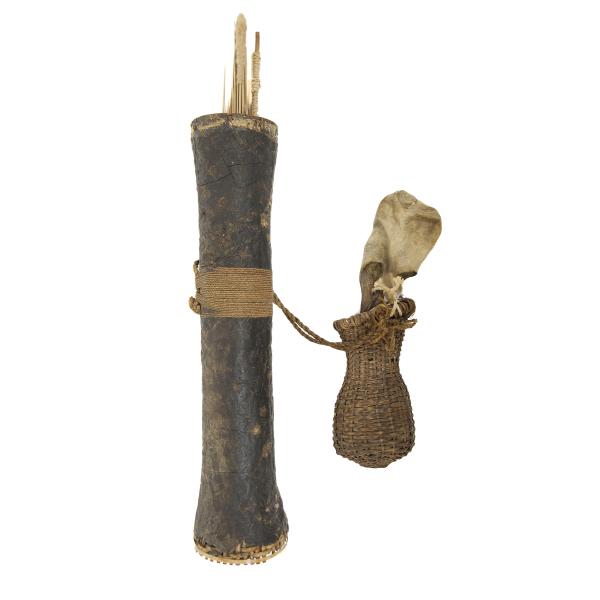 A woven reed quiver covered in cracked black rubber. There are light brown darts coming out of the top. A small woven basket with a skin bag outside of it is attached to the quiver with rope that wraps around the middle of the quiver.