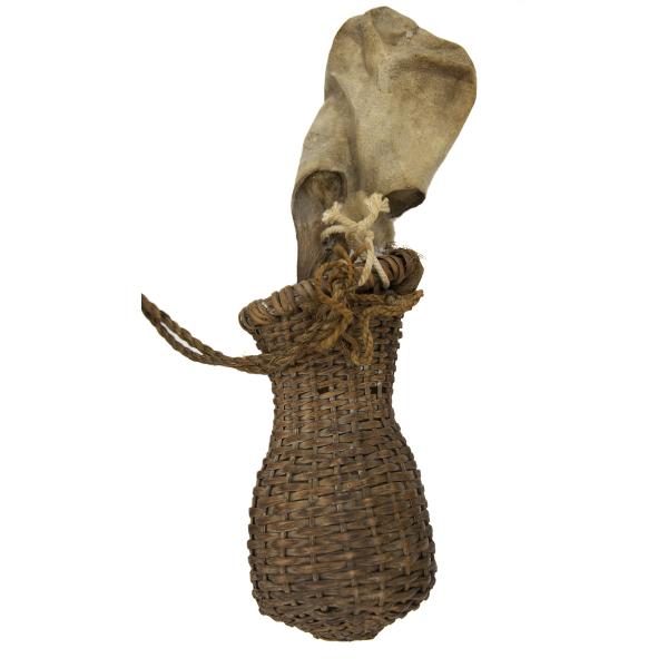 A brown woven reed basket that is tapered and then flares out at the top. There is a skin back sticking out of it that is attached with brown rope.