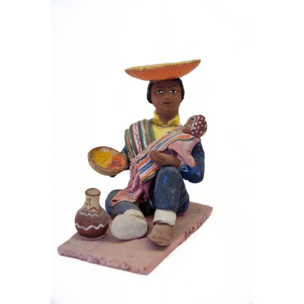 Une figurine montant un homme assis sur un tapis rose et nourrissant un bébé. Il porte un chapeau inversé orange, des sandales, des pantalons bleus, une chemise bleue, une veste jaune, ainsi qu’un poncho vert drapé sur une épaule. Il y a un vase à côté de son pied droit.