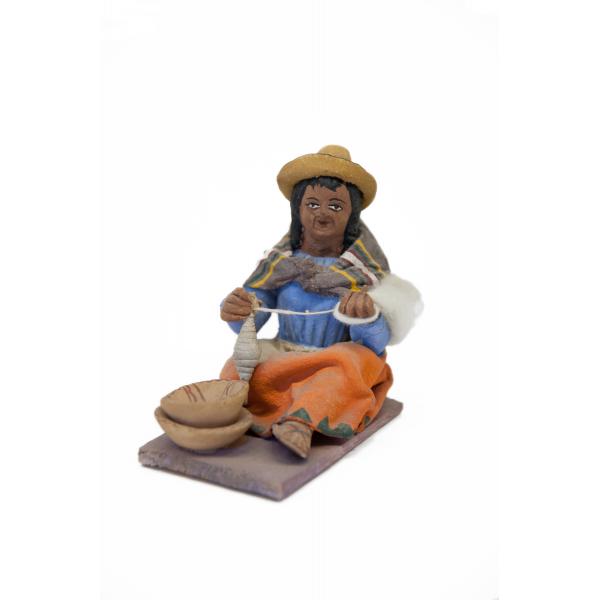 Figurine d’une femme filant de la laine, vêtue de sandales, d’une jupe orange, d’un tablier blanc, d’un chemisier bleu et d’un châle rectangulaire brun. Elle porte un chapeau jaune et deux bols bruns sont posés à ses pieds. 