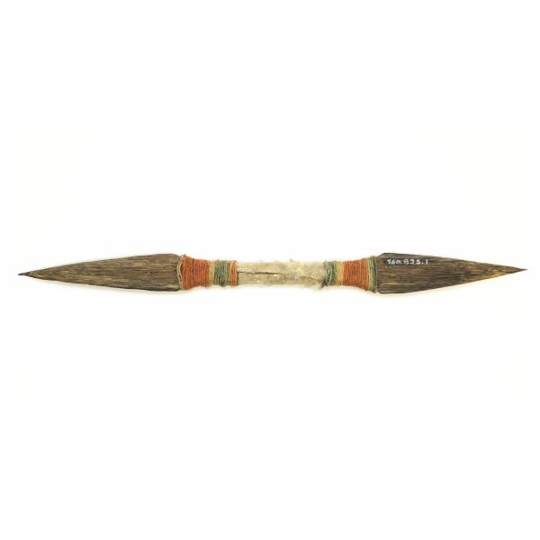 The back of a double-ended wooden knife toy. It is dark brown with points on either end. There is white fur wrapped around the centre. Green, orange, and natural-coloured twined are wrapped around either side of the fur before the points. 