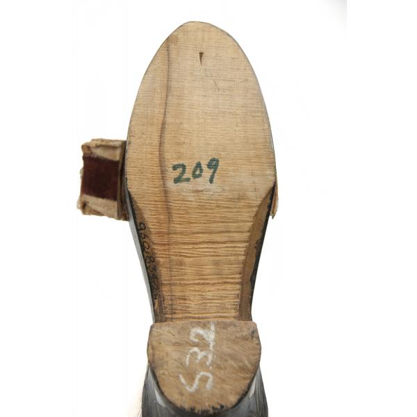 The bottom of a wooden shoe. It is light brown in colour. The heel has been painted black and the fabric strap is sticking out to the left side. 