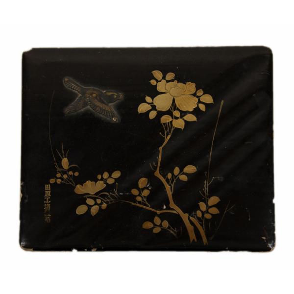 The top of a black box decorated with a gold flower and a metal bird. There are Japanese characters in the bottom left-hand corner beside the flower. 