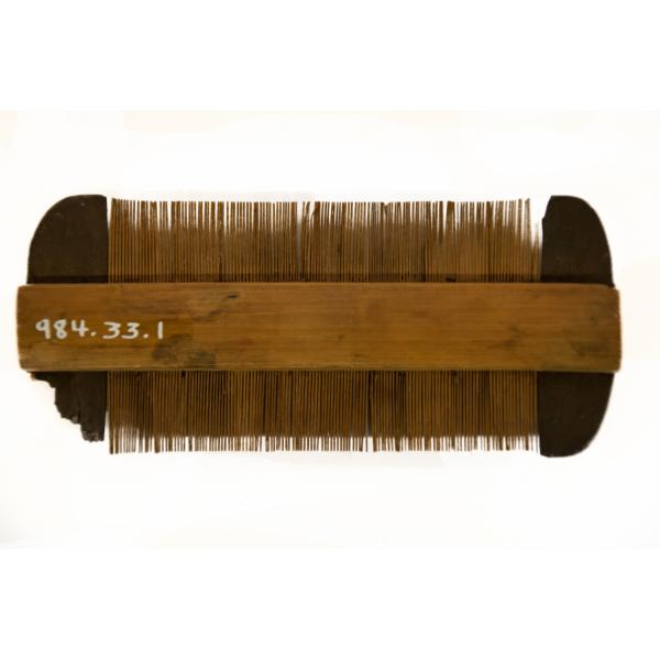 The back of a wooden comb with Japanese characters on the shaft. The teeth are perpendicular to the shaft and are the same brown colour. There is a wider piece of dark brown material at each end. 